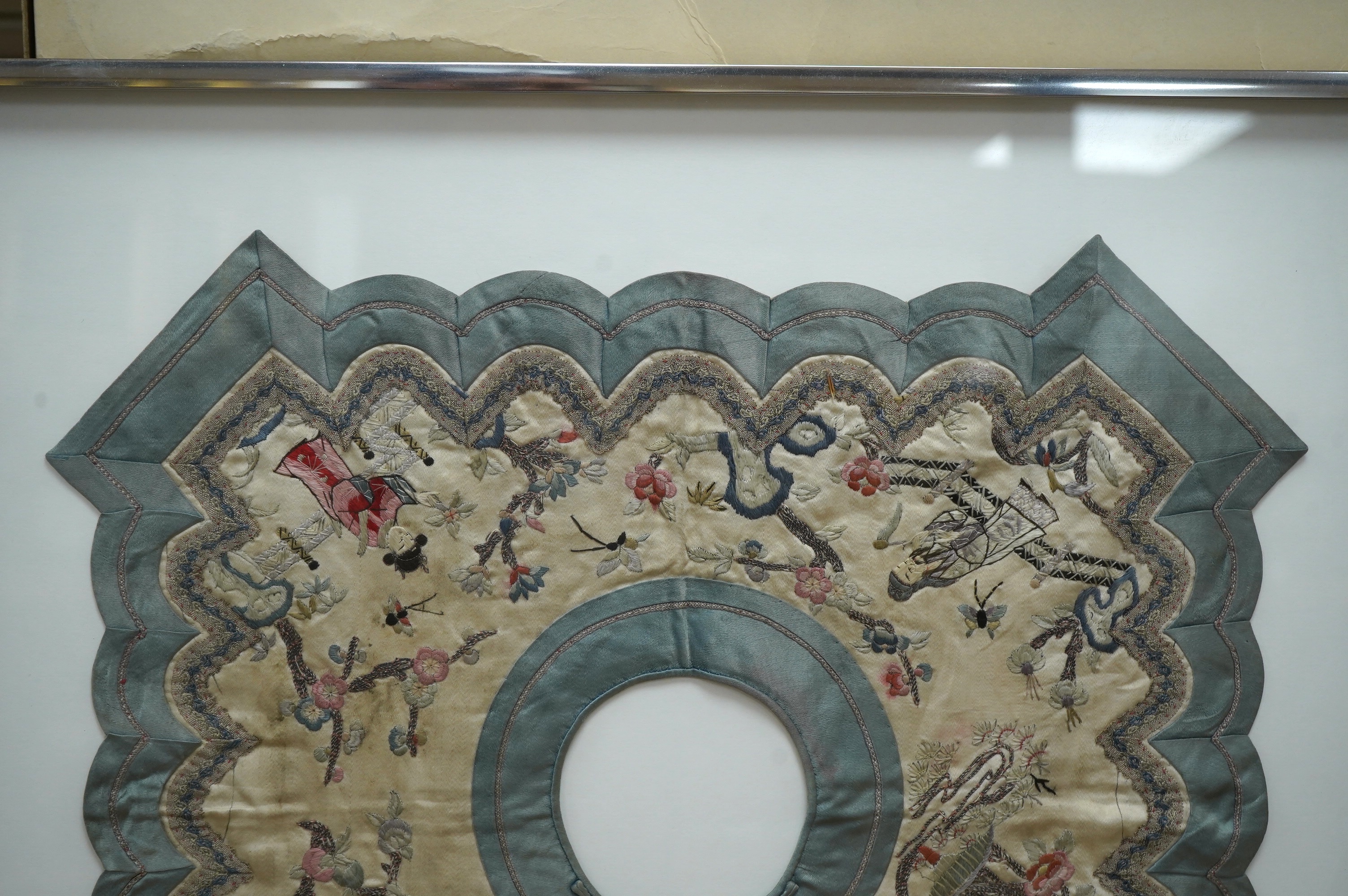 A framed Chinese ornate embroidered collar with scalloped edge, embroidered on cream silk, with polychrome silks, of figures, blossom, butterflies and auspicious symbols, the edges scalloped and braided with an outer blu